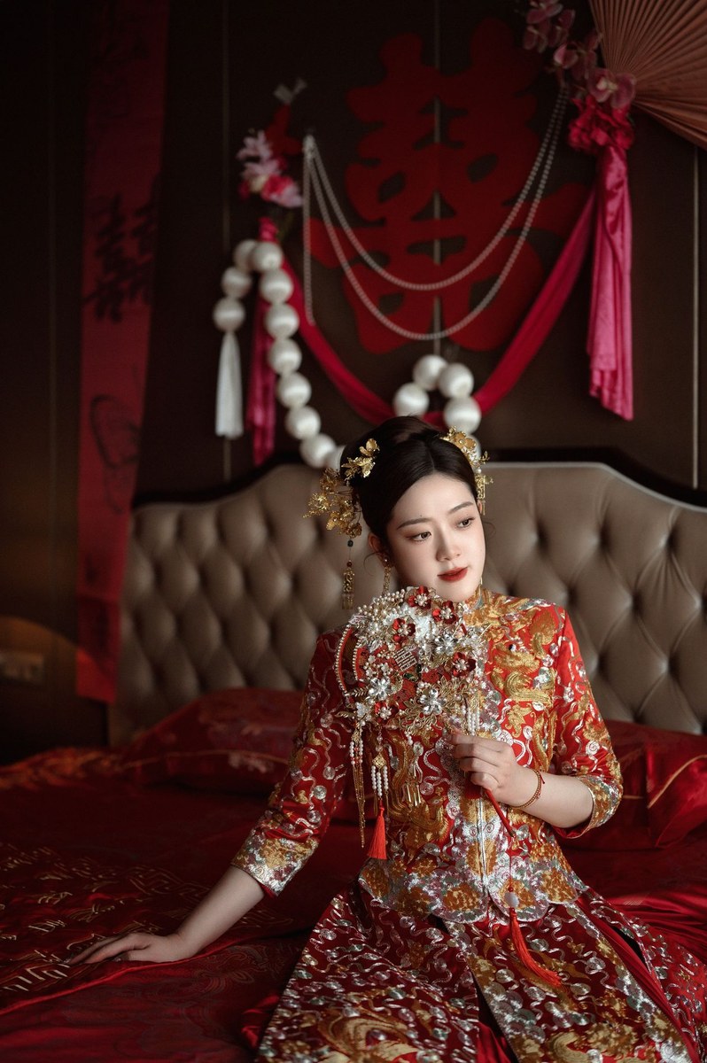 Bride in a red embroidered qun kua holding an ornate pearl bridal fan while seated on red silk bedding