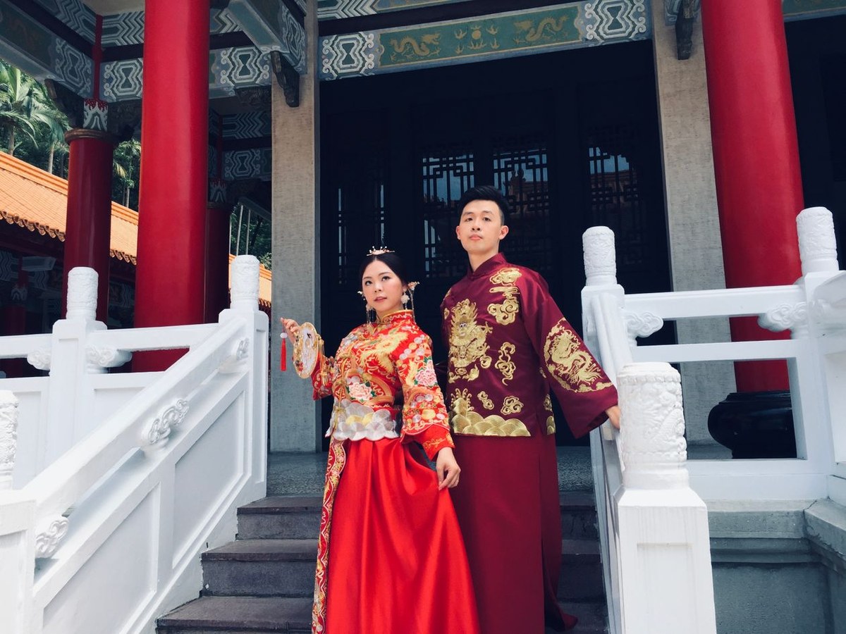 Couple in red-and-gold traditional Chinese wedding attire posing on temple stone steps between red pillars